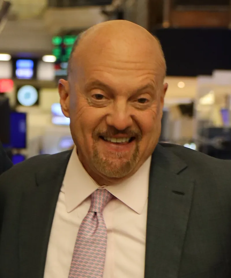Jim Cramer smiling on the floor of the New York Stock Exchange in a dark suit, pale pink shirt and pink patterned tie, with trading screens blurred behind him.